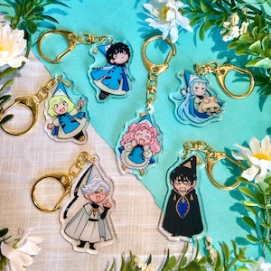 Könnte beinhalten: Sechs bunte Schlüsselanhänger mit Cartoon-Charakteren, goldenen Ketten und Schlüsselringen. Die Figuren tragen blaue, weiße und schwarze Outfits mit spitzen Hüten. Die Schlüsselanhänger befinden sich auf einem blauen und weißen Hintergrund.