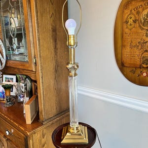 May include: A tall, elegant table lamp with a clear, fluted glass column and a square, gold-toned base. The lamp features a gold-toned metal top and a white lightbulb. It stands on a small, round, dark brown table. The lamp is approximately 30 inches tall.