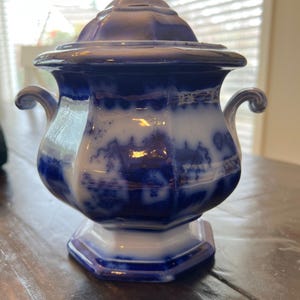 May include: A blue and white ceramic jar with a lid. The jar has a bulbous body, two curved handles, and a hexagonal base. The lid is domed and topped with a small finial. The jar's design features a blue and white pattern.