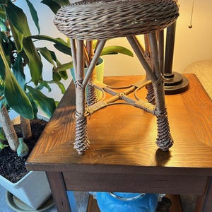 May include: A small, round, woven wicker stool with four legs. The stool has a natural brown color and is placed on a wooden table. The legs are connected with woven supports. The stool is a decorative piece.