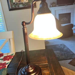 May include: A bronze-colored desk lamp with a bell-shaped, frosted glass shade. The lamp has a round base and a curved arm. The lamp is on a dark wooden table. The lamp is turned on, emitting a warm glow.