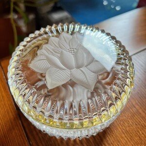 May include: A clear glass trinket box with a cut glass design. The lid features a raised, frosted flower design.