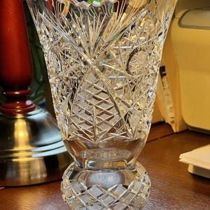 May include: A clear, cut-glass vase with a wide, scalloped rim. The vase features intricate geometric patterns, including diamond and starburst designs. The base is a rounded shape with a patterned design. The vase is approximately 8 inches tall.