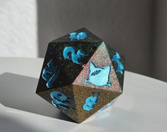 May include: A dodecahedron die with a black and gold glitter finish and blue symbols. The 12-sided die is designed for tabletop games. The blue symbols are stylized and add a unique aesthetic. The die is likely made of resin or a similar material.