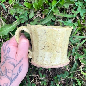 May include: A handmade ceramic mug with a light yellow, speckled glaze and a textured, unglazed reddish-brown base. The mug features a curved handle with a small decorative swirl at the bottom and an uneven, organic rim shape.