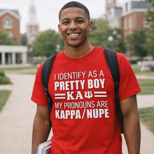 May include: Red t-shirt with white text that reads "I IDENTIFY AS A PRETTY BOY =KAΨ= MY PRONOUNS ARE KAPPA/NUPE." The shirt is worn by a person with a black backpack, walking on a path.