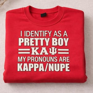 May include: A folded red sweatshirt with white text that reads "I IDENTIFY AS A PRETTY BOY =KA&Psi;= MY PRONOUNS ARE KAPPA/NUPE". The sweatshirt is made of a soft material.