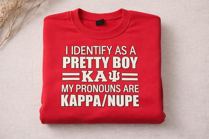 May include: A folded red sweatshirt with white text that reads "I IDENTIFY AS A PRETTY BOY =KA&Psi;= MY PRONOUNS ARE KAPPA/NUPE". The sweatshirt is made of a soft material.