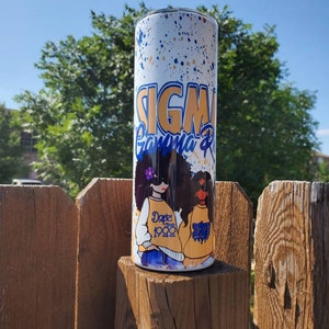 May include: A white tumbler with a blue and gold design featuring the text "Sigma Gamma Rho" and two women with the text "Dope Since 1922".