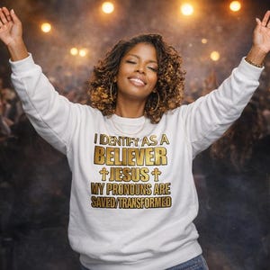 Pode incluir: Uma camisola branca com texto dourado que diz "I IDENTIFY AS A BELIEVER + JESUS + MY PRONOUNS ARE SAVED/TRANSFORMED". A camisola é usada por uma pessoa com os braços levantados. O fundo está desfocado com luzes.