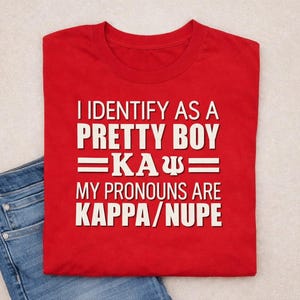 May include: A red t-shirt with white text that reads "I IDENTIFY AS A PRETTY BOY =KA&Psi;= MY PRONOUNS ARE KAPPA/NUPE." The shirt is folded and placed on top of a pair of blue jeans.