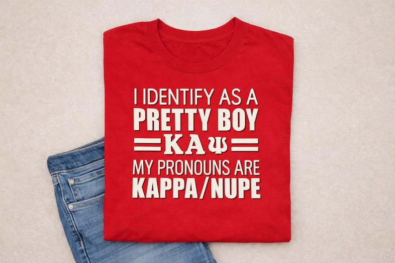 May include: A red t-shirt with white text that reads "I IDENTIFY AS A PRETTY BOY =KA&Psi;= MY PRONOUNS ARE KAPPA/NUPE." The shirt is folded and placed on top of a pair of blue jeans.