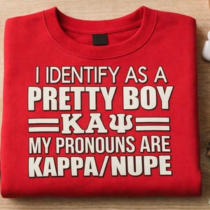 May include: A red sweatshirt with the text "I IDENTIFY AS A PRETTY BOY KAPPA/NUPE" in white block letters. The sweatshirt is folded neatly on a wooden surface, with accessories like glasses, money, and a notebook nearby.