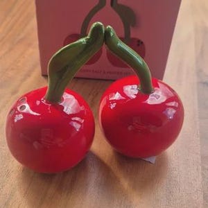May include: Two glossy red cherry-shaped salt and pepper shakers with green stems. They are in front of a pink box with the words "sass & belle" and a cherry design. The set is on a wooden surface.