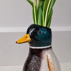 May include: A ceramic mallard duck vase featuring a glossy finish with a dark green head, a bright yellow beak, a white neck ring, and a brown and white body. The vase is filled with several vibrant purple tulips with green stems and leaves.