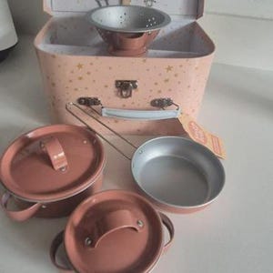 May include: A pink toy kitchen set with a suitcase-style carrying case. The case is open, revealing utensils. Pots, pans, and a colander are arranged in front. The set is designed for children.