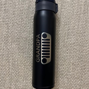 May include: A black insulated water bottle with a dark blue lid. The bottle features the word "GRANDPA" and a silver Jeep grill design. The bottle is designed to keep beverages cold or hot.