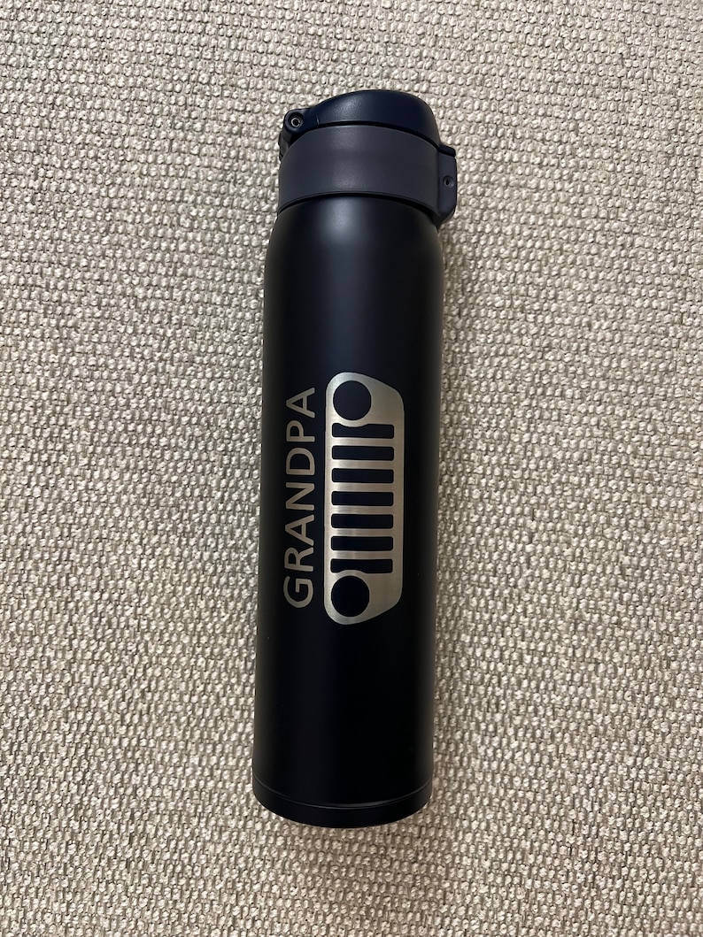 May include: A black insulated water bottle with a dark blue lid. The bottle features the word "GRANDPA" and a silver Jeep grill design. The bottle is designed to keep beverages cold or hot.