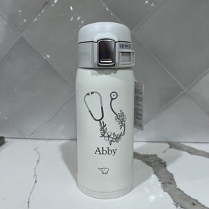 May include: White insulated water bottle with a gray lid. The bottle features a stethoscope design with floral accents and the name "Abby" printed in black. A small bear graphic is also visible.