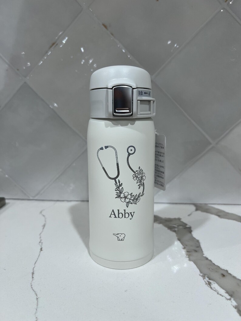 May include: White insulated water bottle with a gray lid. The bottle features a stethoscope design with floral accents and the name "Abby" printed in black. A small bear graphic is also visible.