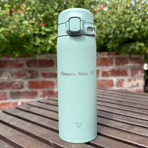 May include: A light green insulated water bottle with a flip-top lid. The bottle has the text "Miranda Metz, MD" printed on it. The bottle is sitting on a wooden surface with a brick wall and greenery in the background.