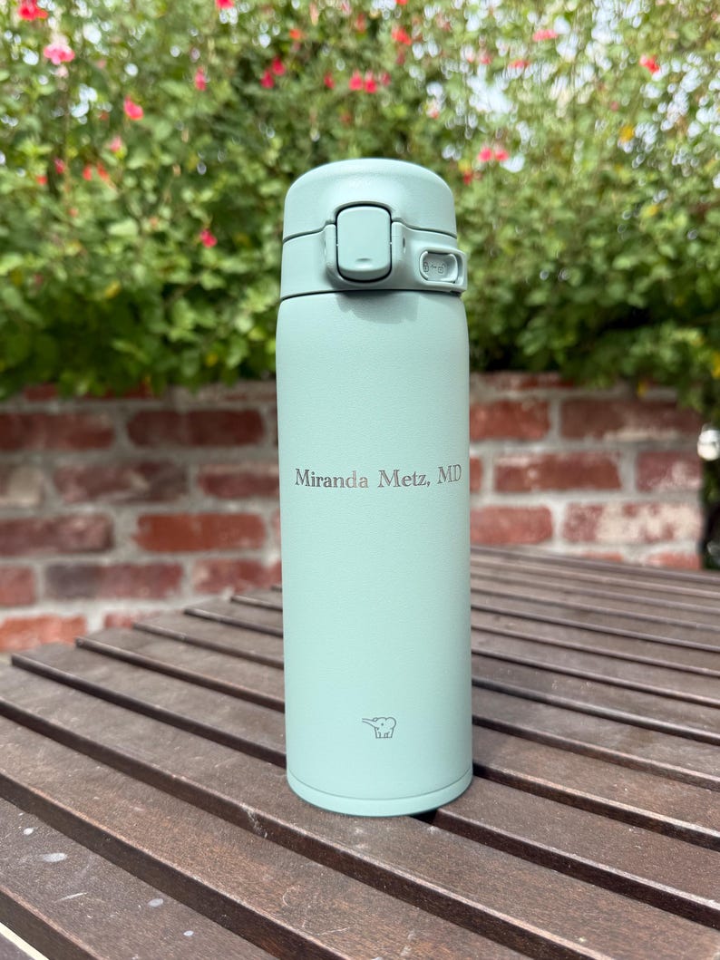 May include: A light green insulated water bottle with a flip-top lid. The bottle has the text "Miranda Metz, MD" printed on it. The bottle is sitting on a wooden surface with a brick wall and greenery in the background.