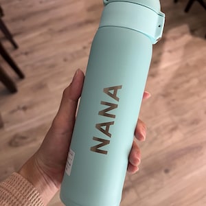 May include: A light blue insulated water bottle with a matching lid. The word "NANA" is printed vertically in a metallic color on the bottle. The bottle is cylindrical and appears to be made of metal.