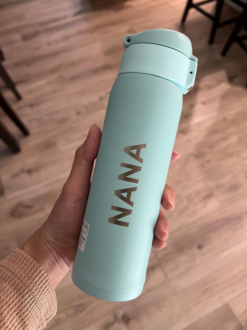 May include: A light blue insulated water bottle with a matching lid. The word "NANA" is printed vertically in a metallic color on the bottle. The bottle is cylindrical and appears to be made of metal.