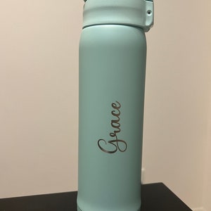 May include: A light blue insulated water bottle with a flip-top lid. The word "Grace" is written in a cursive font on the side of the bottle. The bottle is cylindrical and appears to be made of metal. The bottle is approximately 9 inches tall.