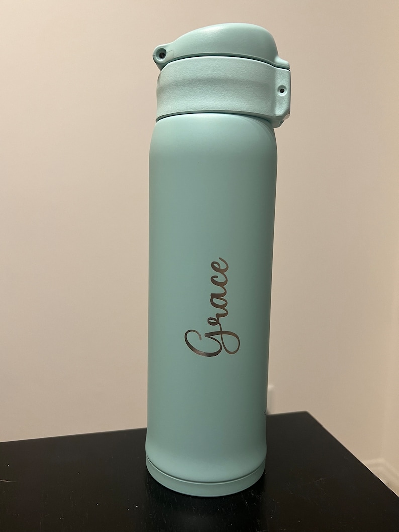 May include: A light blue insulated water bottle with a flip-top lid. The word "Grace" is written in a cursive font on the side of the bottle. The bottle is cylindrical and appears to be made of metal. The bottle is approximately 9 inches tall.