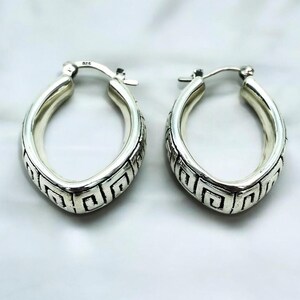 May include: A pair of silver hoop earrings with a geometric design. The earrings are oval shaped and have a Greek key pattern.