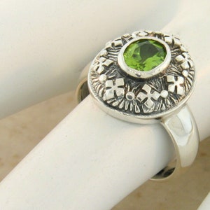 May include: A silver ring with a green gemstone set in a detailed, ornate design. The ring is on a white display stand.