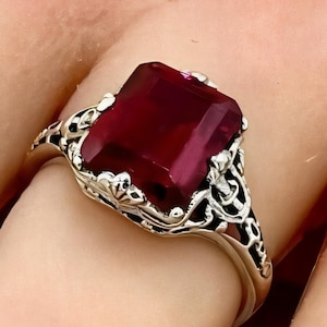 May include: A silver ring with an ornate filigree design features a large, rectangular, deep red gemstone.