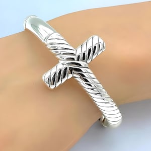 May include: Silver bangle bracelet with a twisted cross design. The bracelet is open and has a smooth, polished finish.