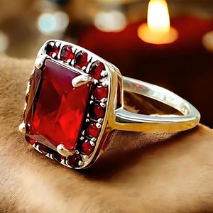 May include: A silver ring with a large, square-shaped, red gemstone surrounded by smaller red gemstones.