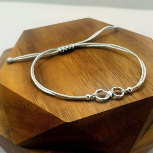 May include: A silver infinity symbol charm bracelet with a white cord. The bracelet is adjustable and has a sliding knot.