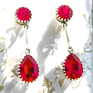 May include: A pair of dangle earrings with red teardrop-shaped stones and smaller round red stones at the top. The earrings are made of gold metal.
