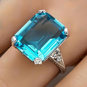 May include: A silver ring with a large, emerald-cut blue topaz gemstone. The ring has a delicate, filigree design.