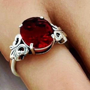 May include: A silver ring featuring a large, oval-cut, deep red gemstone. The ring has an ornate, filigree design on the sides, adding a vintage or antique aesthetic. The gemstone is the focal point, catching the light and drawing attention.