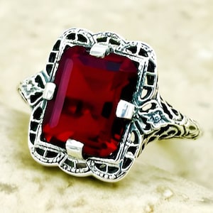 May include: A silver ring with a large, red, square-shaped gemstone set in a filigree design.