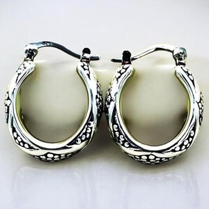 May include: A pair of silver hoop earrings with an ornate, textured design. The earrings have a detailed pattern of small dots and curved lines, creating a vintage or antique aesthetic. The hoops are oval-shaped and feature a secure clasp.