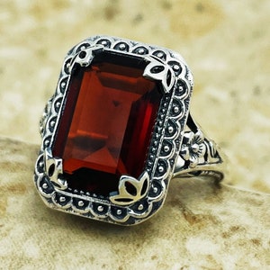 May include: A silver ring with an ornate design featuring a large, rectangular, dark red gemstone.