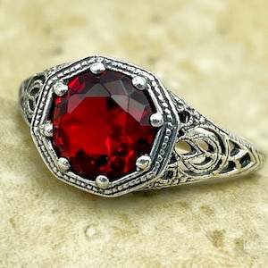 May include: A silver ring with an ornate filigree design and a large, red gemstone in the center.