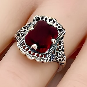 May include: A silver ring with an ornate filigree design. The ring features a large, rectangular, deep red gemstone. The setting is detailed with openwork patterns, creating an antique or vintage aesthetic. The ring is shown on a person's finger.