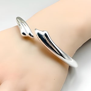 May include: A silver bangle bracelet with a unique, curved design. The bracelet is open and has a smooth, polished finish.
