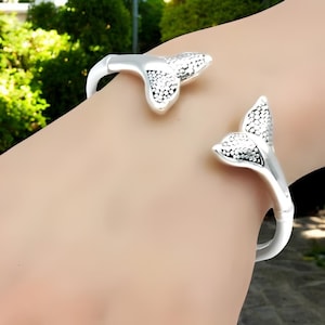 May include: A silver-colored cuff bracelet with a mermaid tail design. The bracelet features two stylized tail fins with a textured, dotted pattern. The bracelet is open, allowing it to be slipped onto the wrist.