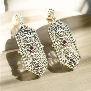 May include: A pair of gold filigree earrings with red gemstone accents. The earrings are long and dangle, with a delicate, intricate design.