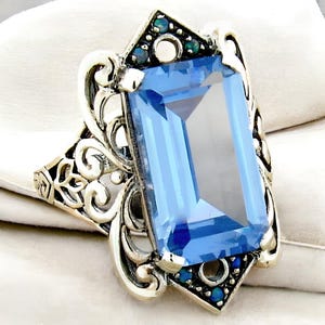 May include: A silver ring with a large, rectangular, light blue gemstone surrounded by a filigree design and smaller, dark blue gemstones.