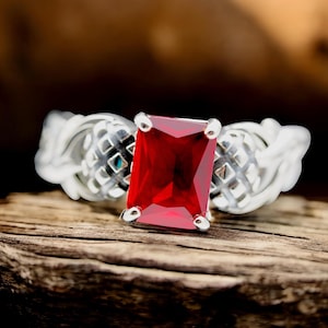 May include: A silver ring with a large, square-shaped, red gemstone set in the center. The ring has a detailed, intricate design.