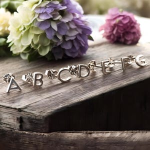 May include: A set of silver initial letter earrings, from A to G, displayed on a wooden surface. The earrings are small and delicate, perfect for adding a touch of personalization to any outfit.